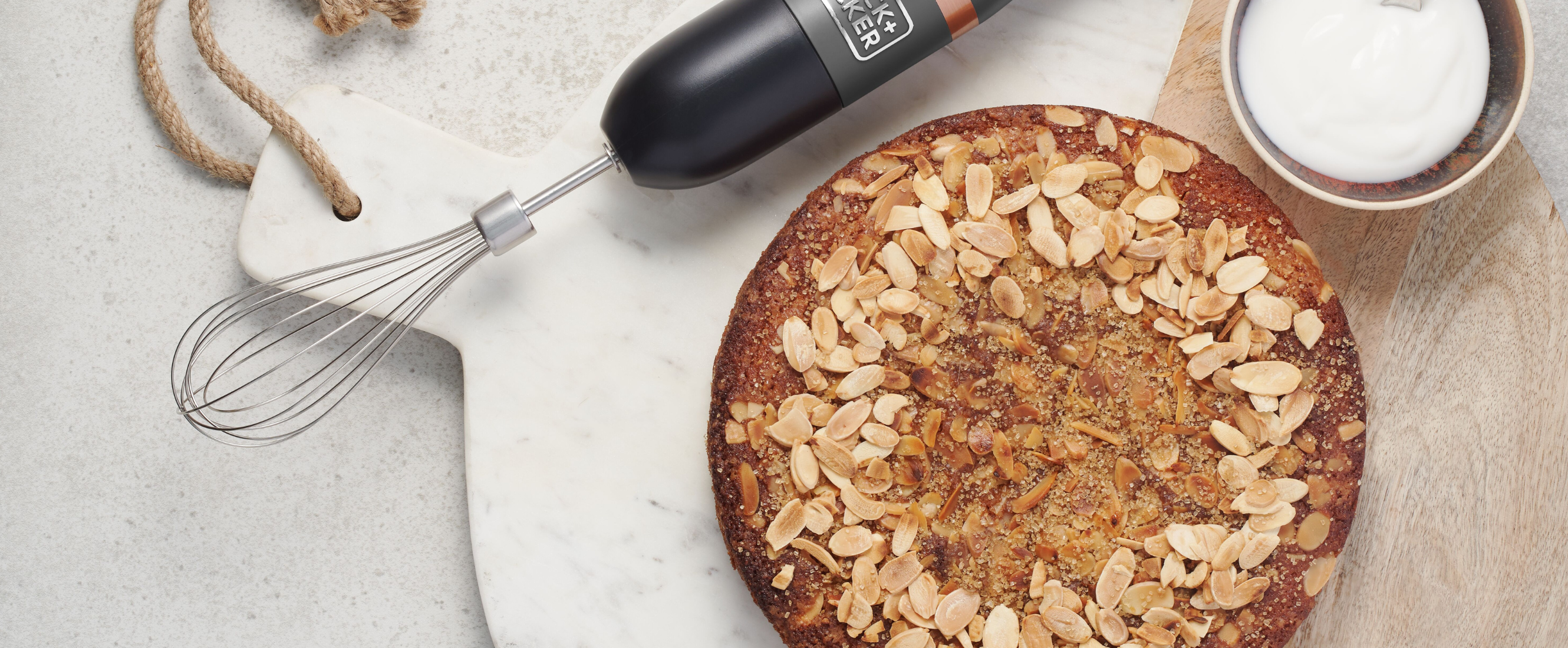 Grey, Black-and-decker kitchen wand with whisk attachment lying next to a marble plate with a gluten free cherry and almond cake