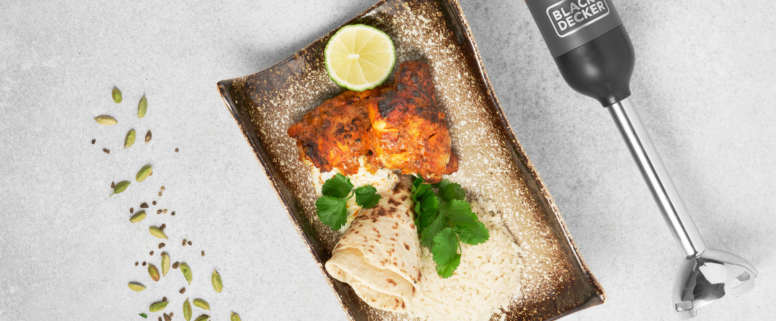 Grey, Black-and-decker kitchen wand with immersion blender attachment lying next to a plate of tandoori chicken