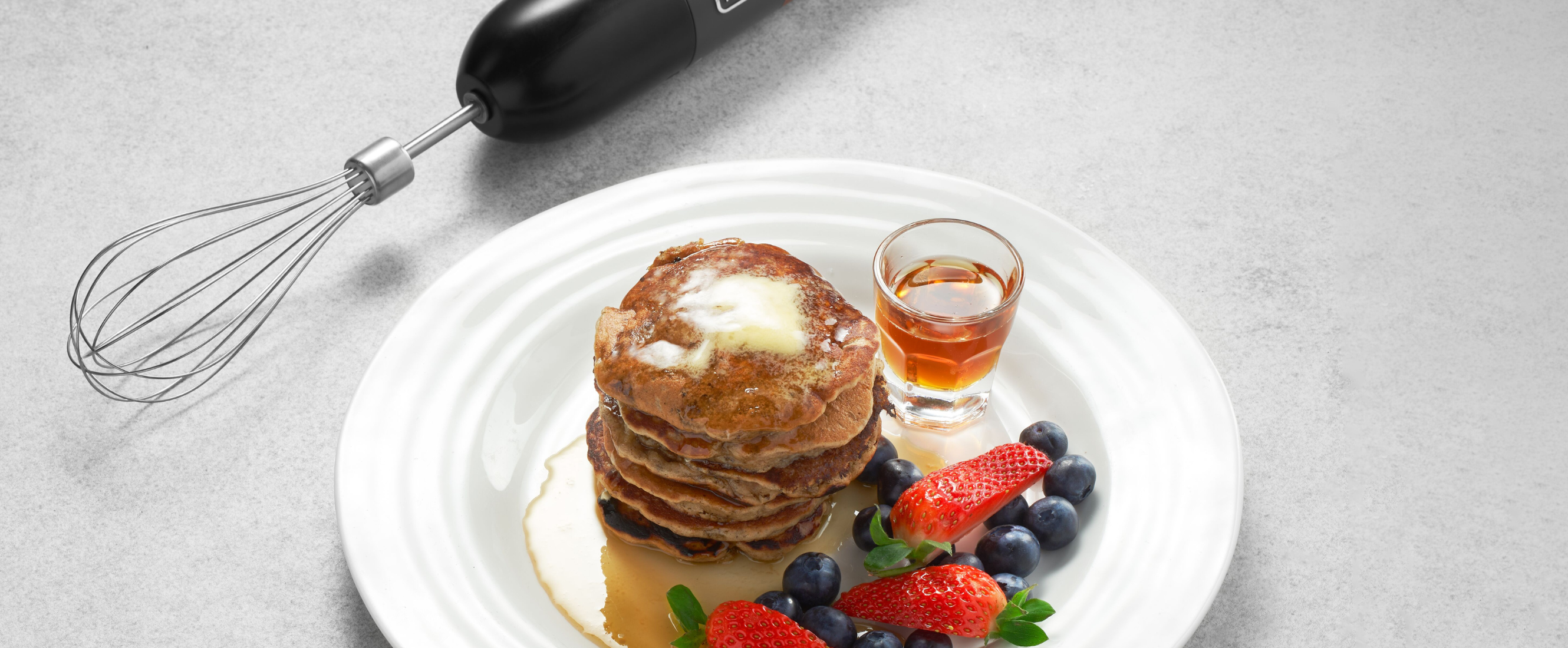 Grey, Black-and-decker kitchen wand with whisk attachment lying next to a plate of pancakes, mixed berries, and maple syrup