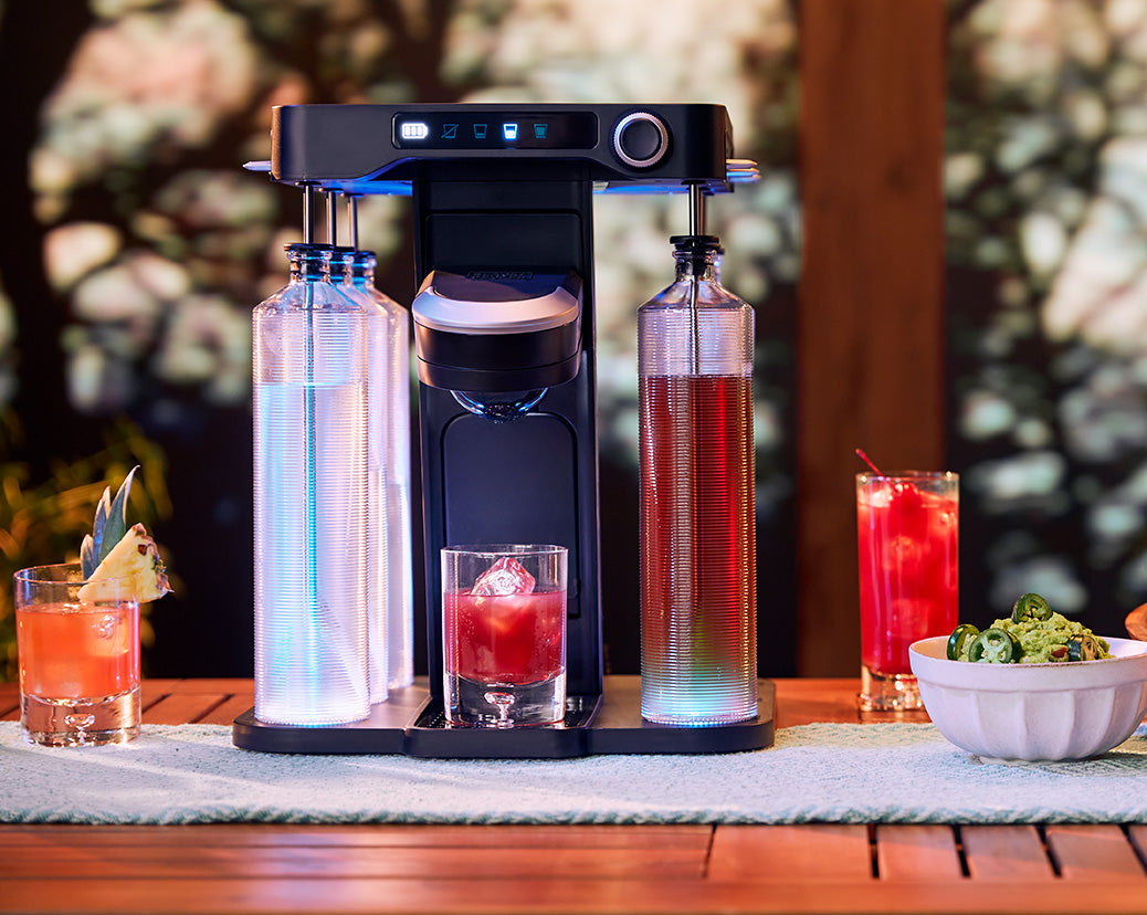 blue lights illuminated on the bev by Black-and-decker™ cordless cocktail maker sitting on a wooden patio table with drinks and appetizers